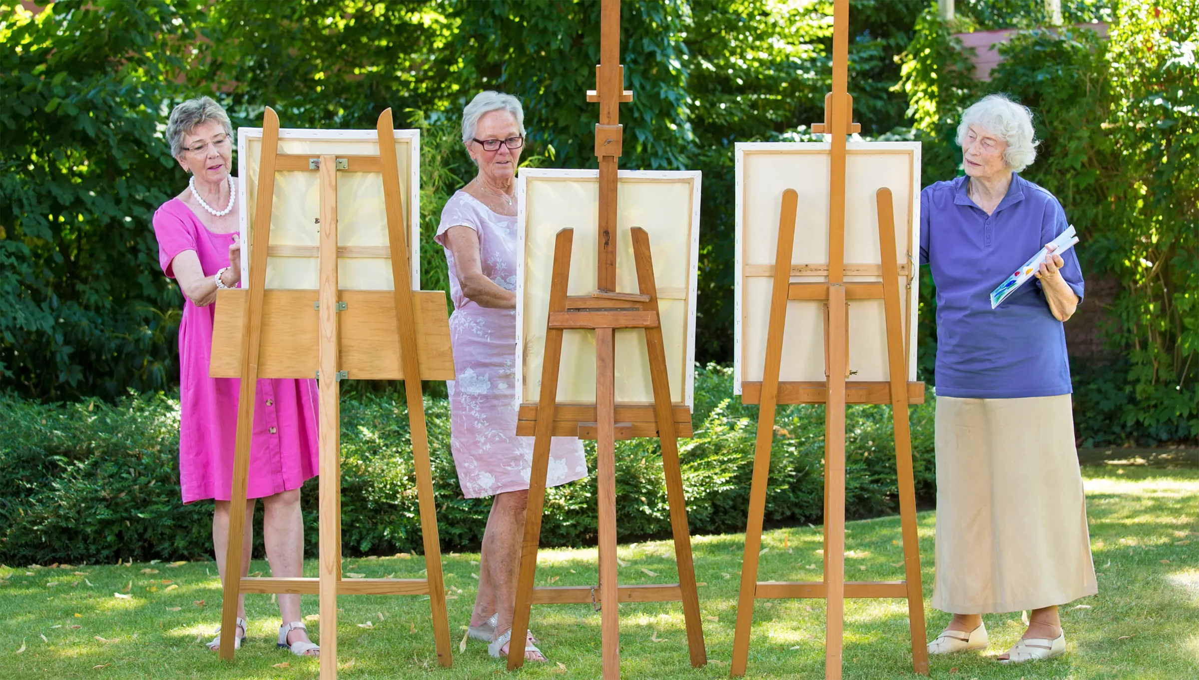 Three female residents standing together outside on the grass painting on easels.